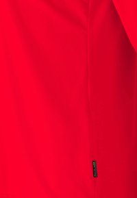Rotes kurzärmliges Oberteil aus glattem, leichtem Stoff. Verfügt über eine Seitennaht und ein kleines Markenetikett nahe dem Saum.