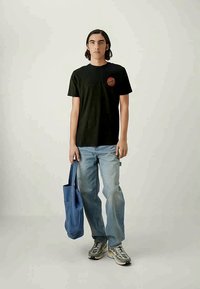 Joven de pie, vistiendo una camiseta negra de Santa Cruz, jeans azul claro, zapatillas grises, sosteniendo una bolsa de tela azul en la mano derecha.