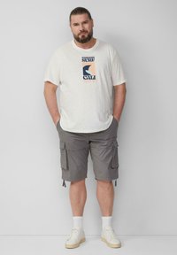 Mann mit Bart, der ein weißes Grafik-T-Shirt, graue Cargo-Shorts, weiße Socken und weiße Sneaker trägt und vor einer schlichten grauen Wand steht.