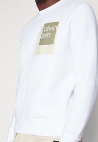 Sudadera de cuello redondo blanca con un logo gráfico en oliva y beige. Fabricada con tejido suave y con puños y dobladillo acanalados.