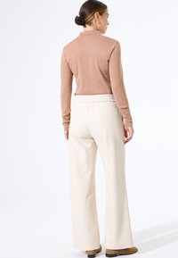 Langsleeve, lichtbruine top met hoge kraag en een aangesloten silhouet; gecombineerd met losse, crèmekleurige broeken en bruine schoenen.