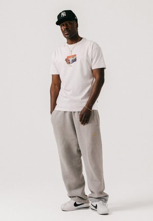 Hombre con gorra de béisbol negra, camiseta blanca con gráfico, pantalones deportivos grises y zapatillas Nike blancas, de pie frente a un fondo claro y sencillo.