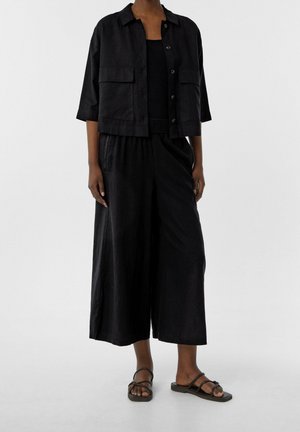 Kobieta ubrana w czarną, krótko skrojoną kurtkę na guziki z kieszeniami, spodnie o szerokich nogawkach do połowy łydki oraz czarne, płaskie sandały z paskami, stojąca na szarej podłodze.