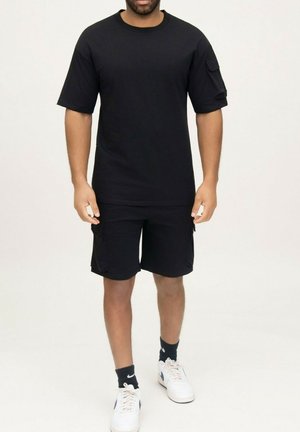 Homme portant un t-shirt noir à manches courtes avec une poche, un short cargo noir, des chaussettes Nike noires et des baskets Nike blanches, debout sur fond blanc.