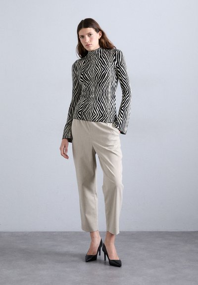 Haut à col roulé rayé noir et blanc avec une coupe ajustée, associé à un pantalon beige clair sur mesure et des talons noirs, sur un fond gris.