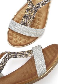 Zwei Sandalen mit einer strukturierten goldenen Innensohle und silbernen geflochtenen Riemen. Ein Riemen weist ein geflochtenes Muster mit goldenen und grauen Akzenten auf.