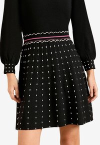 Černé plisované šaty se vzorem bílých čtverců, žebrovaným pasem s růžovými a bílými akcenty a dlouhými nafouknutými rukávy.