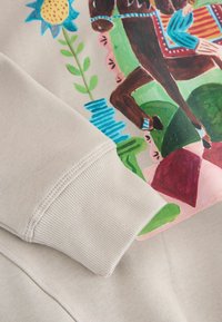 Beige pulóver színes illusztrációval, amelyen egy ló, növények és geometriai formák láthatók, bordázott mandzsettákkal és sima textúrával.