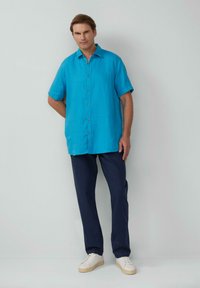Lichtblauwe linnen overhemd met korte mouwen en knoopsluiting, gecombineerd met marineblauwe broek en witte sneakers, staand tegen een neutrale achtergrond.