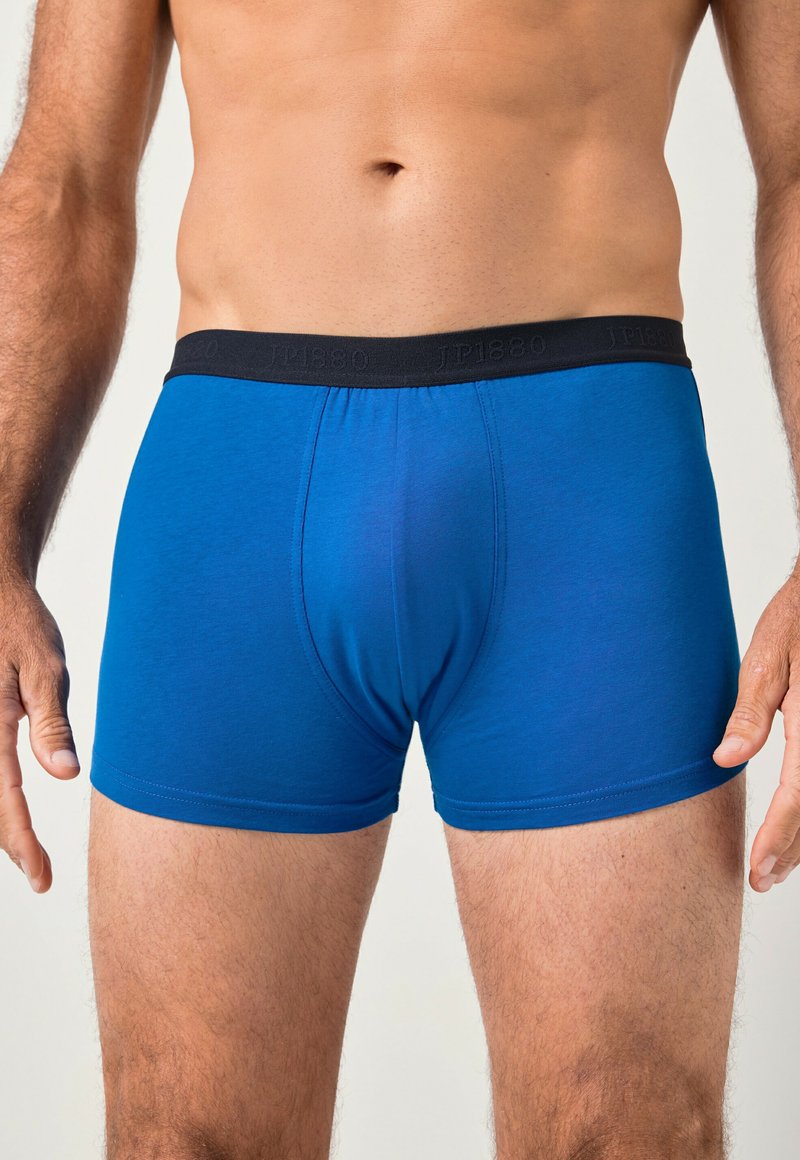 Männlicher Oberkörper, der blaue Boxershorts mit einem schwarzen Bund und schwacher Markenkennzeichnung trägt, vor einem schlichten hellen Hintergrund stehend.