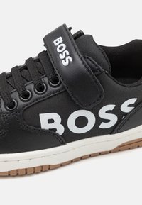 Schwarzer Sneaker mit schwarzen Schnürsenkeln, weißen "BOSS"-Logos an der Seite und am Riemen, weißer Zwischensohle und brauner strukturierter Außensohle.