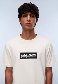 Cremefarvet t-shirt lavet af bomuld. Har et sort rektangulært logo med teksten "NAPAPIJRI" og "geographic" i hvid og sort. Kortærmet.