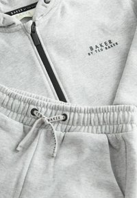 Sudadera gris con cremallera y pantalones de chándal a juego, hechos de una tela suave, con cremallera negra y cordones, y detalles de logo en ambas prendas.