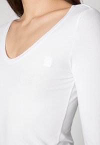 Vit långärmad topp gjord av slätt tyg med djup V-ringning och en subtil logodetalj på bröstet. Enkel, figurnära design.