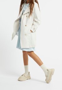 Jasny trencz z złotymi guzikami, zestawiony z niebieską plisowaną sukienką i kremowymi masywnymi mokasynami, stylizowany z podkolanówkami.