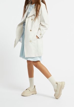 Trench ligero de color claro con botones dorados, combinado con un vestido azul plisado y mocasines gruesos color crema, estilizado con calcetines hasta la rodilla.