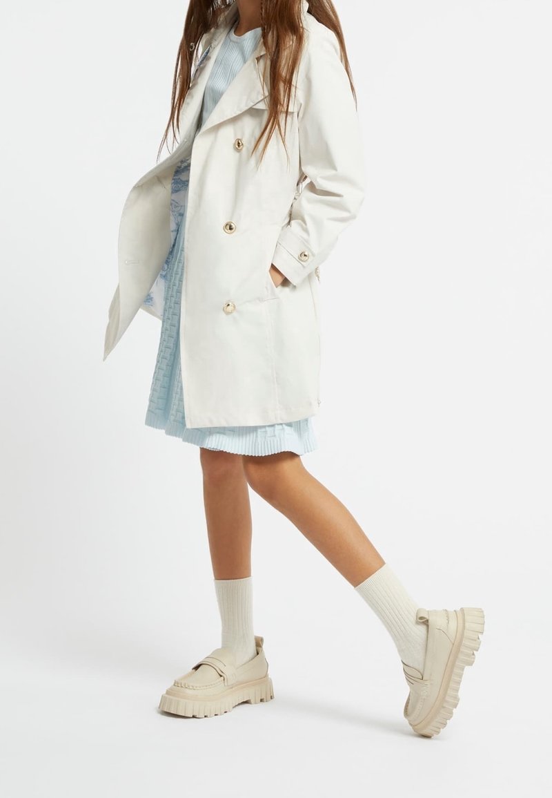 Jasny trencz z złotymi guzikami, zestawiony z niebieską plisowaną sukienką i kremowymi masywnymi mokasynami, stylizowany z podkolanówkami.