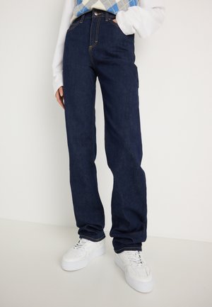Persona che indossa jeans a gamba dritta blu scuro, una maglietta bianca a maniche lunghe e sneaker bianche con zeppa, in piedi contro uno sfondo neutro.
