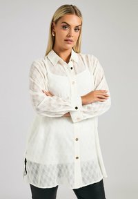 Camicia bianca con bottoni, con un motivo testurizzato, maniche lunghe e bottoni dorati. Il tessuto è leggero e leggermente trasparente.