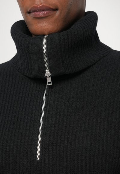 Must mustriba turtleneck-sweater, millel poolt tõmbe sulg. Krae on paks ja tekstureeritud. Materjal tundub pehme ja soe.