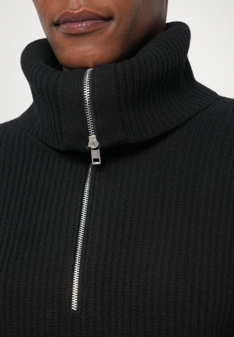 Svart ribbad polo tröja med halvdragkedja och en tjock, texturerad krage. Tyget ser mjukt och varmt ut.