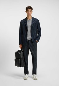 Marinblått rutigt kostym med en enkelknappad kavaj, grå skjorta, svart läderryggsäck och ljusa sneakers, med en slät textur och strukturerad design.
