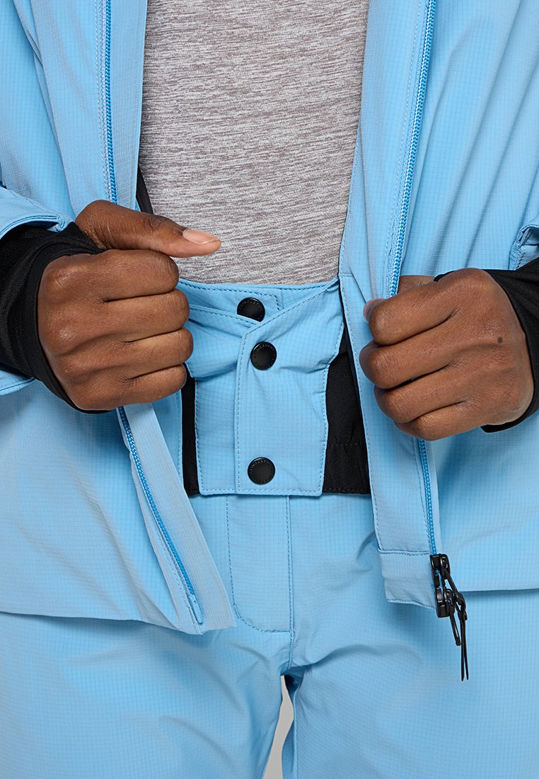 Ręce zapinają zamek błyskawiczny i kolorowe guziki na jasnoniebieskiej kurtce i spodniach na szarym t-shircie, koncentrując się na detalach zapięcia.