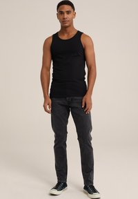 Črna majica brez rokavov, sivi jeans in črne športne copate z belimi podplati; model stoji, nevtralna ozadja poudarja obleko.