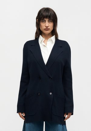 Femme aux cheveux foncés portant un blazer bleu marine croisé, une chemise blanche boutonnée et un jean bleu, debout devant un fond clair uni.
