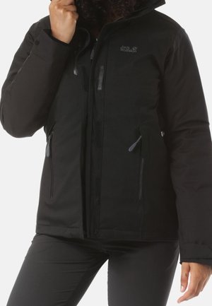 Person trägt eine schwarze Jack Wolfskin Jacke mit Reißverschlusstaschen, richtet den hohen Kragen zurecht und trägt passende schwarze Hosen.