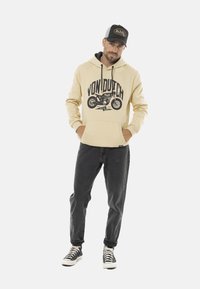 Hoodie beige avec le logo noir "Von Dutch" et un graphique de moto, associé à un jean foncé et des baskets noires, le tout agrémenté d'une casquette contrastante.
