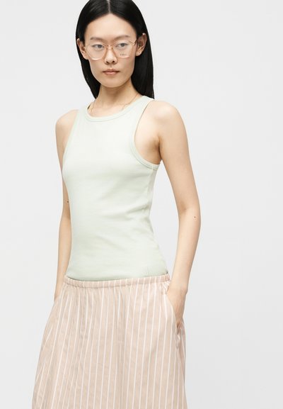 Femme portant des lunettes transparentes couleur pêche, un haut sans manches vert pâle et une jupe beige avec des rayures verticales blanches, debout les mains dans les poches.