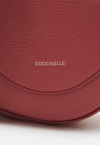 Röd texturerad läderhandväska med en kurvad form, med en subtil präglad logotyp "COCCINELLE" i silver ovanför sömmen.