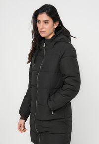 Svart polstret lang kåpe med glidelås, hette og sidelommer; har et teksturert stoff og kontrasterende ribbede mansjetter.