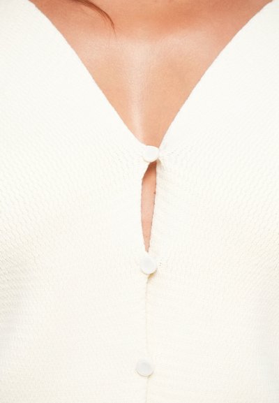 Bela pletena jopica z V-izrezom, okrasno gumbno zapiranje na prsnici in teksturirano blago z rahlim rebrastim vzorcem.