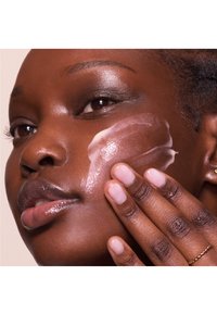 Crema idratante applicata su una guancia; il modello ha la pelle liscia e scura, labbra lucide e smalto per unghie neutro. La pelle ha una texture luminosa.