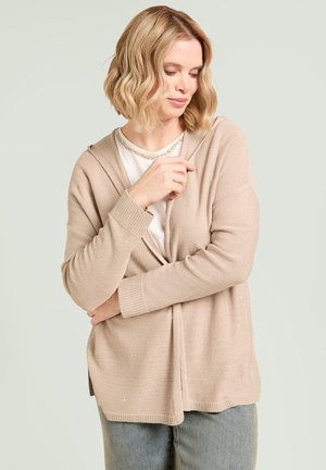 Blond kvinde iført beige strikket cardigan over hvid top og blå jeans, der kigger ned med den ene arm krydset og hånden rører kraven.