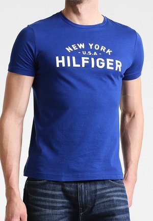 Homme portant un T-shirt bleu à manches courtes avec un texte blanc « New York U.S.A Hilfiger » et un jean bleu foncé sur un fond uni.
