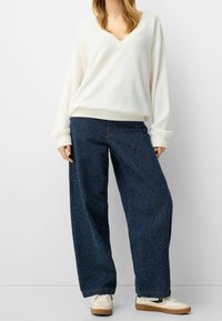 Biały sweter z dekoltem w serek i ściągaczami przy mankietach, zestawiony z luźnymi, ciemnoniebieskimi spodniami dżinsowymi o subtelnej fakturze i swobodnym kroju. Całość dopełniają białe trampki.