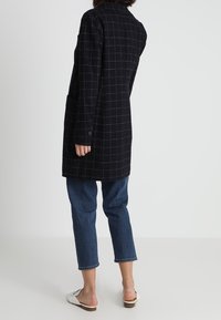 Femme portant un long manteau noir à carreaux, un jean court bleu et des chaussures blanches sans lacets, debout devant un fond clair uni.