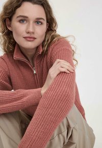 Strickjacke mit Reißverschluss in gedämpftem Pink, aus texturiertem Stoff und mit langen Ärmeln, kombiniert mit hellbeigen Hosen. Modell sitzt, Hände auf den Knien.