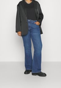 Blazer de satén negro sobre una camisa negra, combinado con unos vaqueros de tiro alto y campana azules y zapatos de plataforma negros. Nota: costuras visibles en los vaqueros.