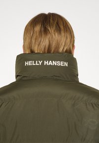 Alyvuogių žalios spalvos pūkinė striukė su aukštu kaklu, ant nugaros užrašyta "HELLY HANSEN" šviesiu tekstu. Lygus paviršius ir aptakus dizainas.
