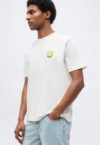 Άνδρας που φοράει λευκό T-shirt με γραφικό ακτινίδιο και κείμενο στο στήθος, ανοιχτόχρωμα μπλε τζιν, στέκεται με το ένα χέρι στην τσέπη.