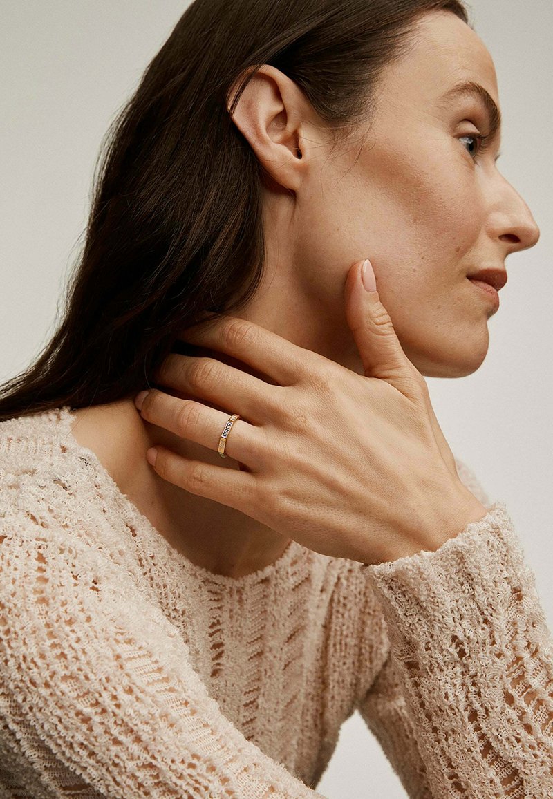 Anillo de oro con una pequeña gema, llevado en una mano con uñas cuidadosamente arregladas. Suave suéter de punto texturizado en color crema. Diseño minimalista.