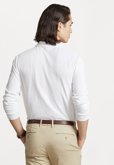 Witte langemouwen polo met kraag, gecombineerd met beige broek en een bruine leren riem. Gladde textuur met minimale details.