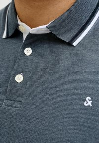 Σκούρο γκρι πόλο μπλουζάκι από υφασμάτινη υφή. Διαθέτει λευκό γιακά με μαύρη ρίγα, τρία λευκά κουμπιά και μικρό λογότυπο στο στήθος.