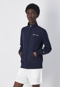 Chaqueta de zip en color navy hecha de tela suave con un cuello alto, que presenta el logo de Champion en el pecho y bolsillos en la parte delantera.