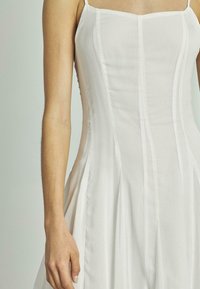 Weißes Kleid mit dünnen Spaghettiträgern, das vertikale Streifen aufweist und einen taillierten Oberteil hat, der in einen plissierten Rock übergeht. Glattes, leichtes Material.