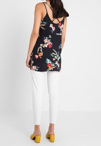 Tunika w kwiatowy wzór z czarnego materiału, z ramiączkami spaghetti i V-kształtnym tyłem, zestawiona z białymi wąskimi spodniami oraz żółtymi sandałami na słupku.